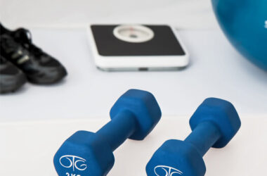 Zwei blaue Kurzhanteln (jeweils 2 kg) liegen auf einer weissen Oberfläche, im Hintergrund sind eine digitale Waage und weitere Fitnessutensilien zu sehen.