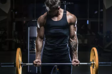Mann hebt eine Langhantel im Fitnessstudio, dabei sind die ausgeprägten Arm- und Schultermuskeln sichtbar, was auf intensives Krafttraining zur Förderung von Muskelwachstum (Hypertrophie) hinweist.