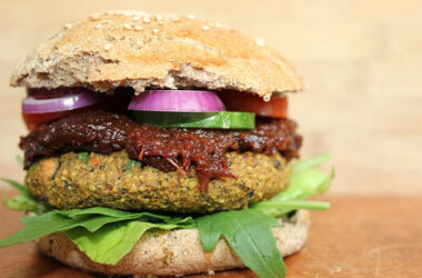 Ein schmackhaft aussehnder veganer Burger mit einem pflanzlichen Patty, frischem Rucola, roten Zwiebeln, Gurkenscheiben, Tomaten und einer würzigen Sauce im Vollkornbrötchen.
