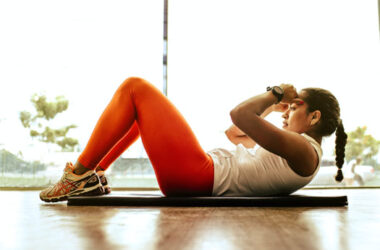 Eine Frau in Sportkleidung macht Sit-ups auf einer Matte in einem lichtdurchfluteten Raum mit grossen Fenstern.