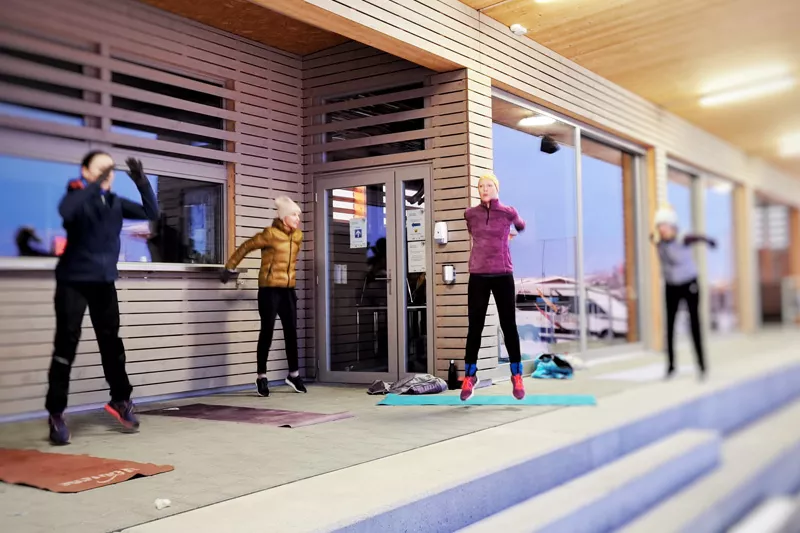 Gruppe von sportlich gekleideten Menschen, die draussen während eines Bootcamp-Training in die Höhe springt, bei kühlem Wetter und vor einem modernen Gebäude.