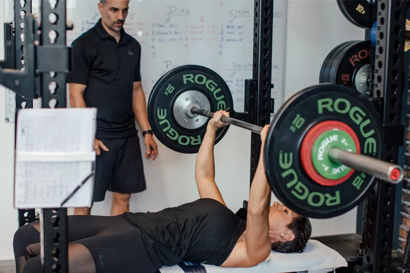 Eine Klientin von Personalworkout führt Bankdrücken mit der Langhantel aus, während ihr Personal Trainer Marc im Hintergrund ihre Technik überwacht, um ihr zu helfen, die Technik zu verbessern und höhere Gewichte zu stemmen.