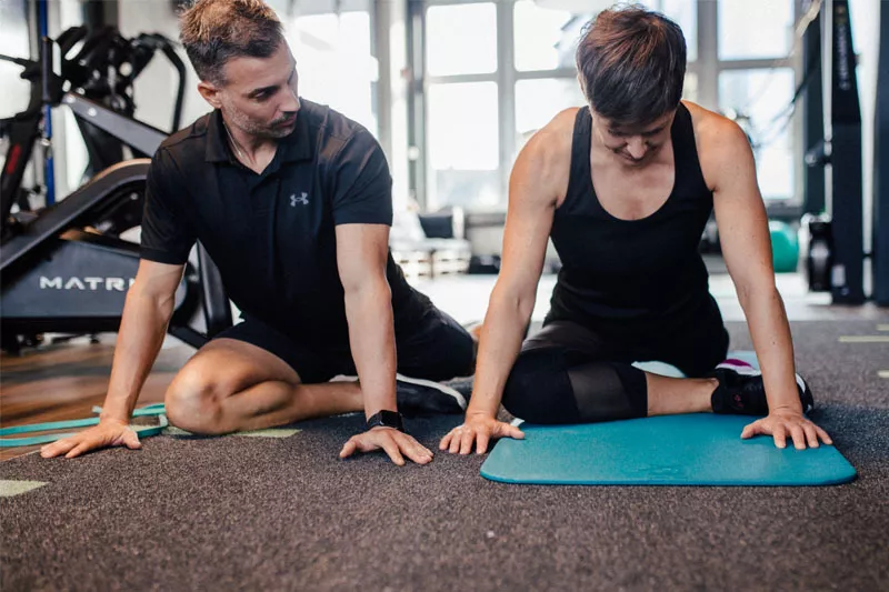 Personal Trainer Marc und eine Klientin führen im Fitnessstudio von Personalworkout gemeinsam den Piriformis Stretch aus, eine Übung, die besonders bei Rückenschmerzen hilfreich sein kann.