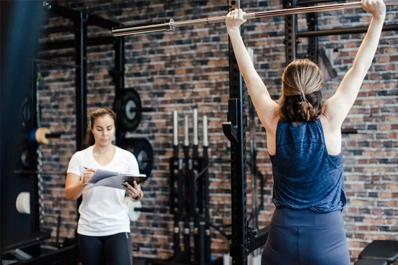 Im modernen FItnessstudio von Personalworkout führt eine Klientin unter Anleitung der Personal Trainerin Sofia eine Military Press mit der Langhantel aus. Die stilvolle Atmosphäre mit Backsteinwänden und hochwertigem Equipment schafft die perfekte Umgebung für effektives Training.