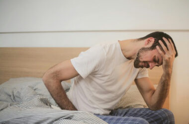Ein Mann in Schlafkleidung sitzt sitzt vornübergebeugt mit schmerzverzerrtem Gesicht im Bett und hält eine Hand an die Seite, während die andere seinen Kopf stützt.