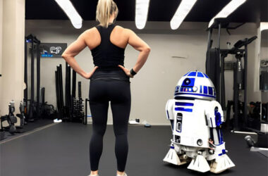Eine Frau in Sportbekleidung steht mit den Händen auf den Hüften in einem Fitnessstudio, während neben ihr ein R2-D2-ähnlicher Roboter auf dem Boden steht. Das Bild spielt humorvoll auf die Frage an, ob KI oder ChatGPT einen Personal Trainer ersetzen können.